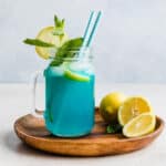 A jar of Blue Raspberry Lemonade on a brown plate, garnished with a sprig of fresh mint and lemon slice.