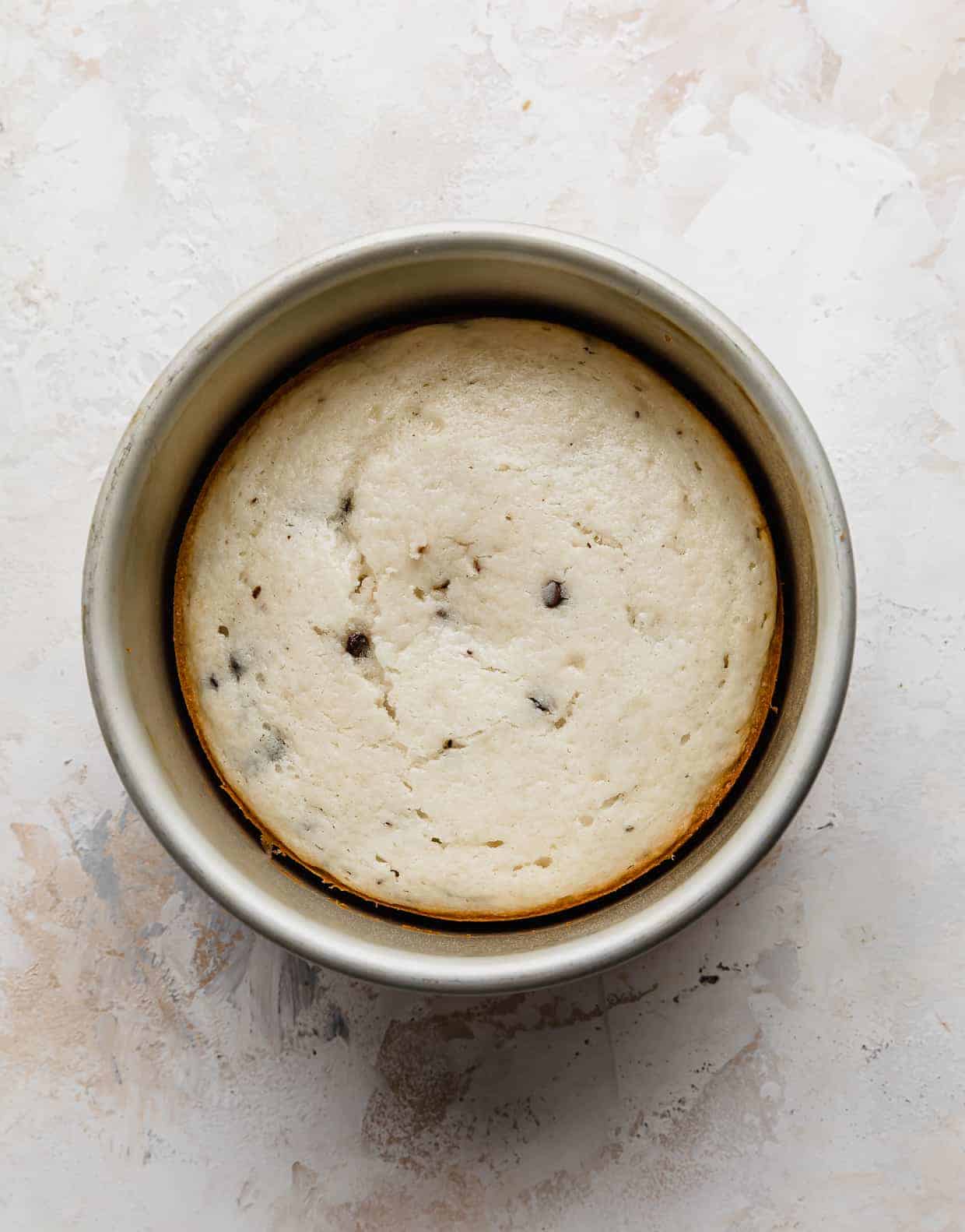A baked white cake with mini chocolate chips in a round cake pan, on a white background.