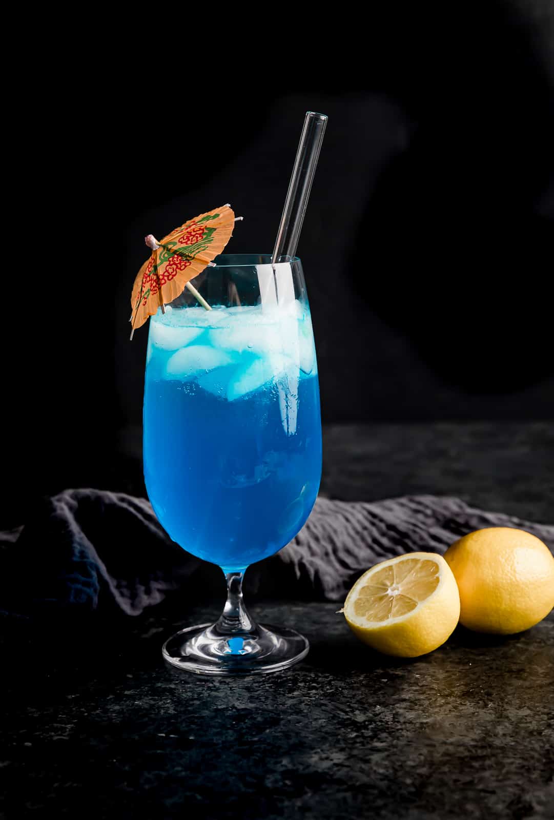 A glass cup full of a blue lagoon drink with a pink umbrella in the cup.