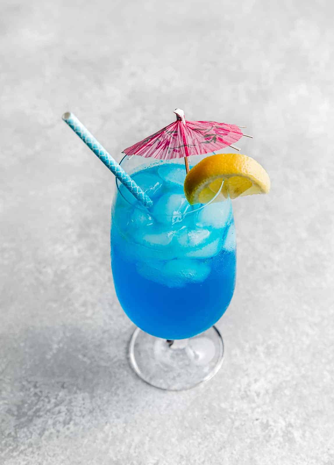 A blue lagoon mocktail drink on a gray background with a pink umbrella and lemon wedge on the edge of the cup.