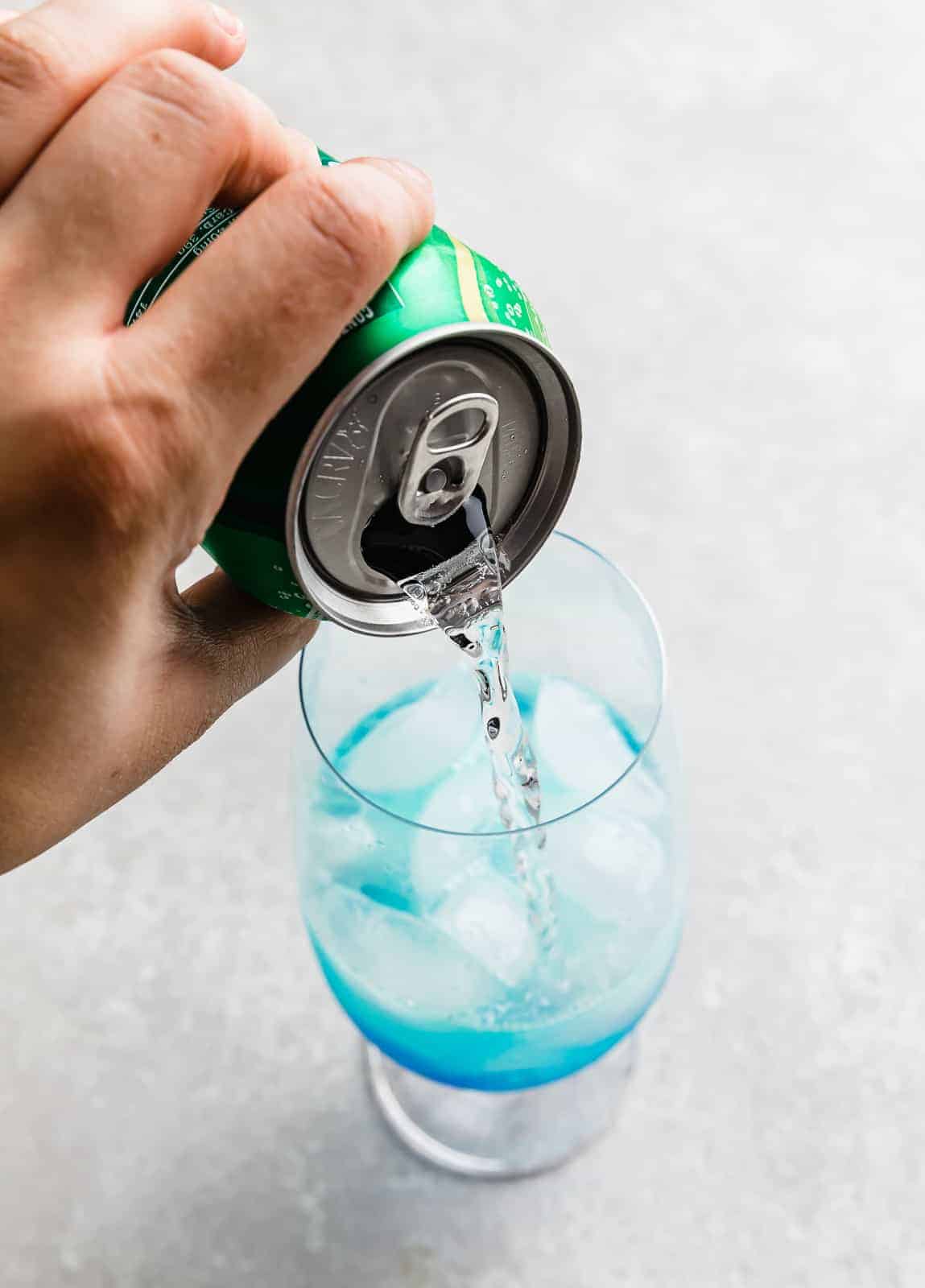 A can of 7 UP being poured into a glass cup that has blue curaçao syrup in it. 