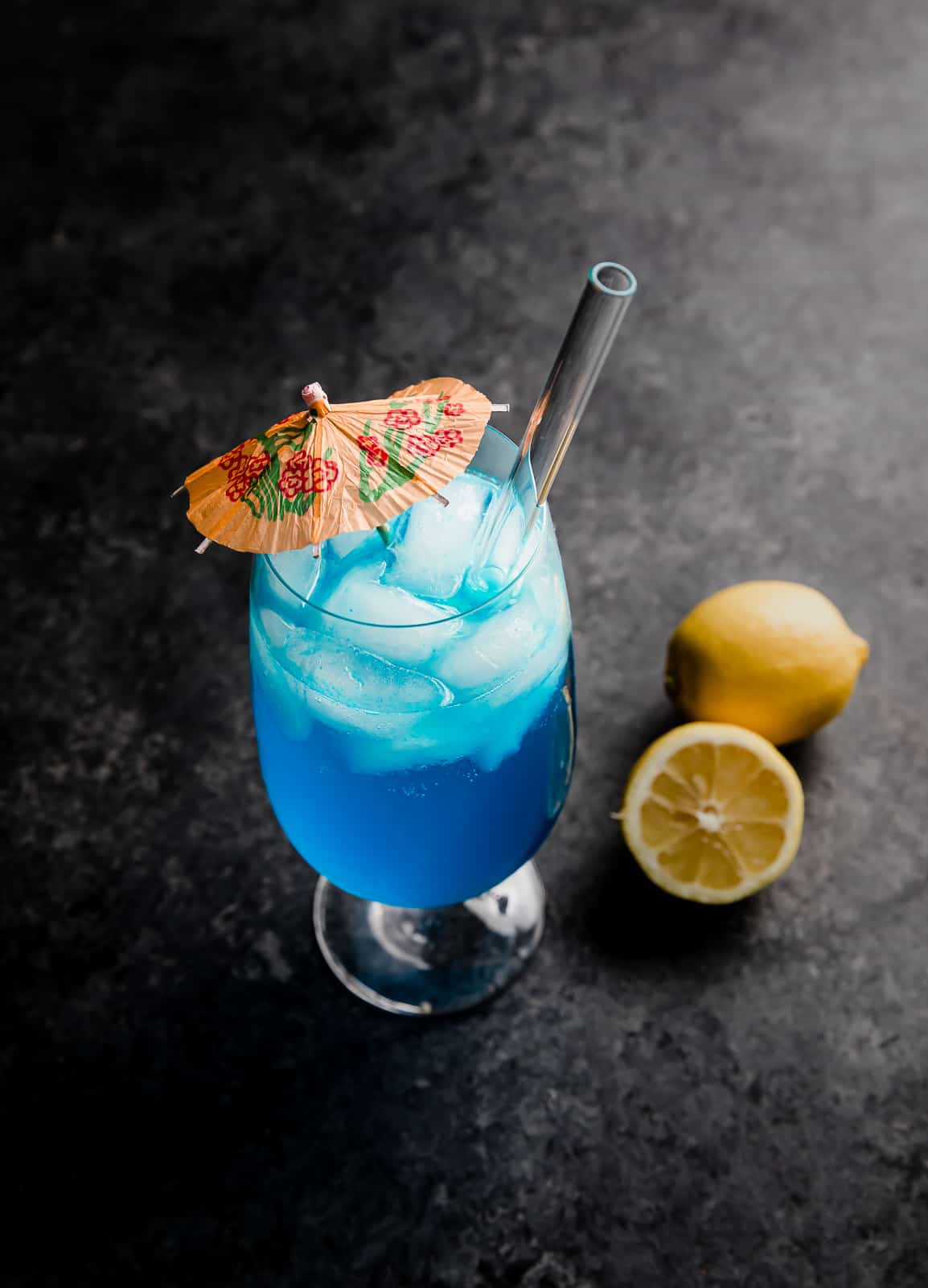 A Blue Lagoon Mocktail on a black background with a lemon next to the glass.