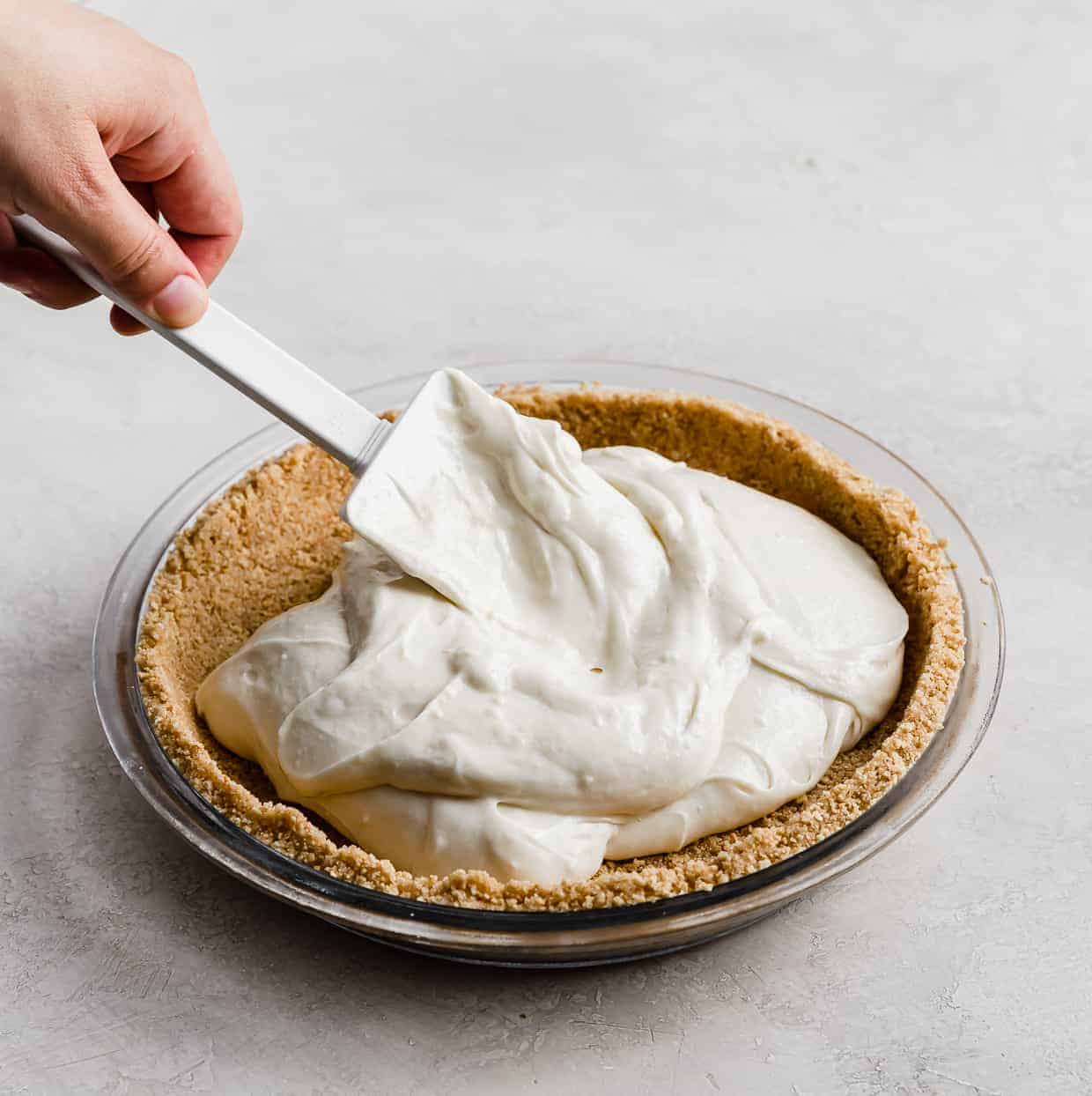 A hand holding a spatula that is spreading the cheesecake filling inside a graham cracker pie crust.