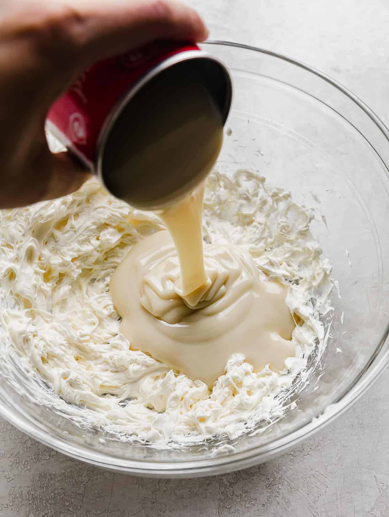 A hand pouring a can of sweetened condensed milk into a glass bowl.