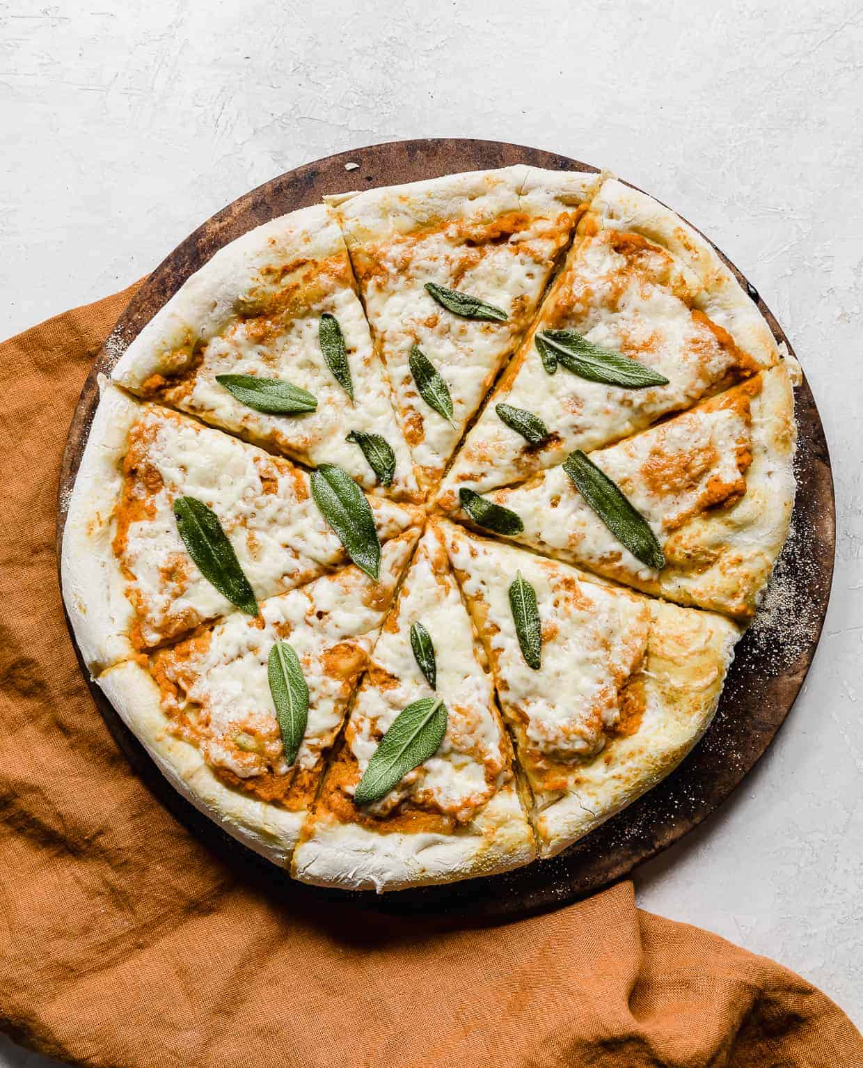 A baked pumpkin pizza topped with sage leaves on a round pizza stone against a gray background.