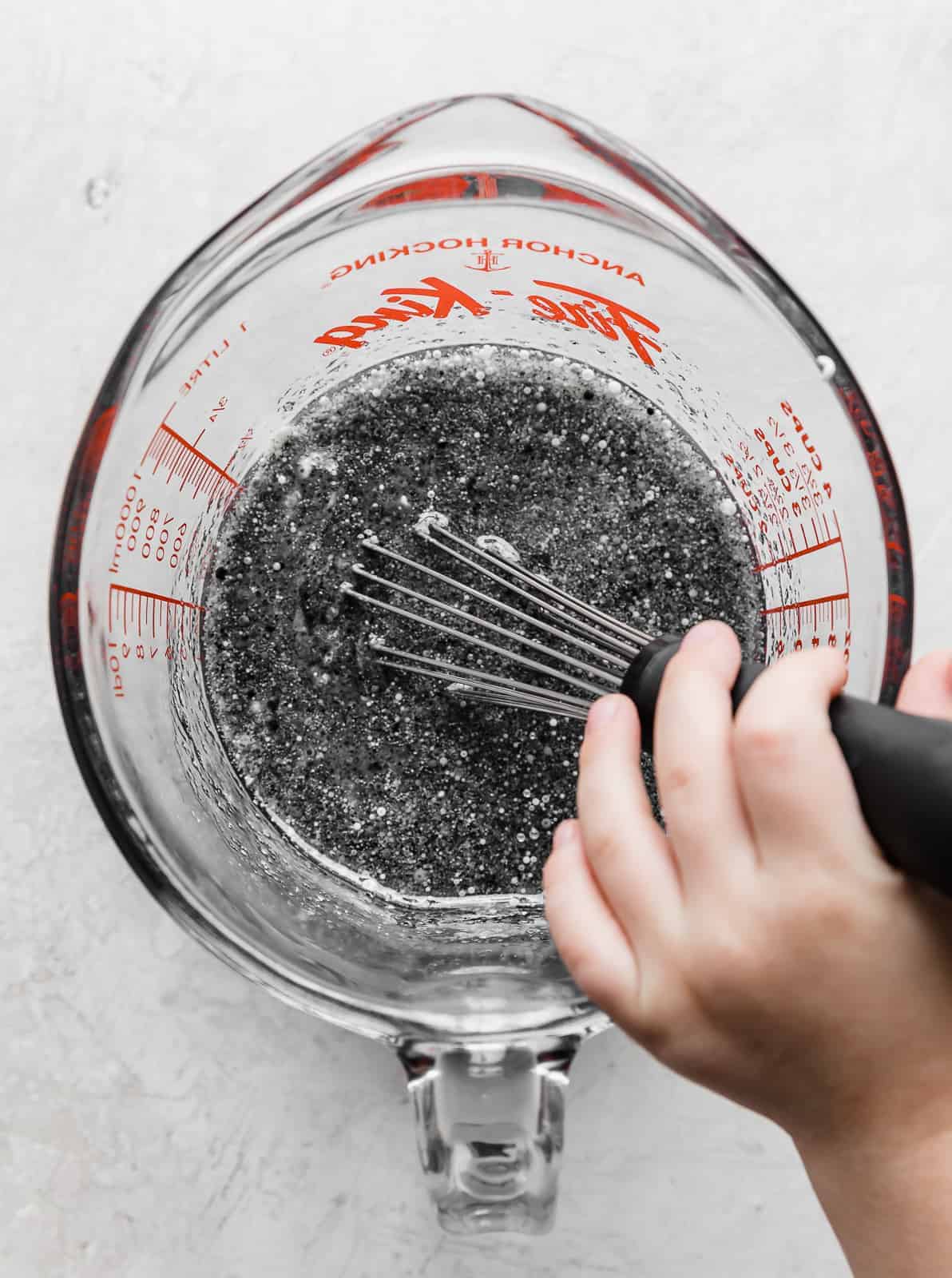 A dark gray liquid mixture in a glass measuring cup, with someone using a whisk to mix it.