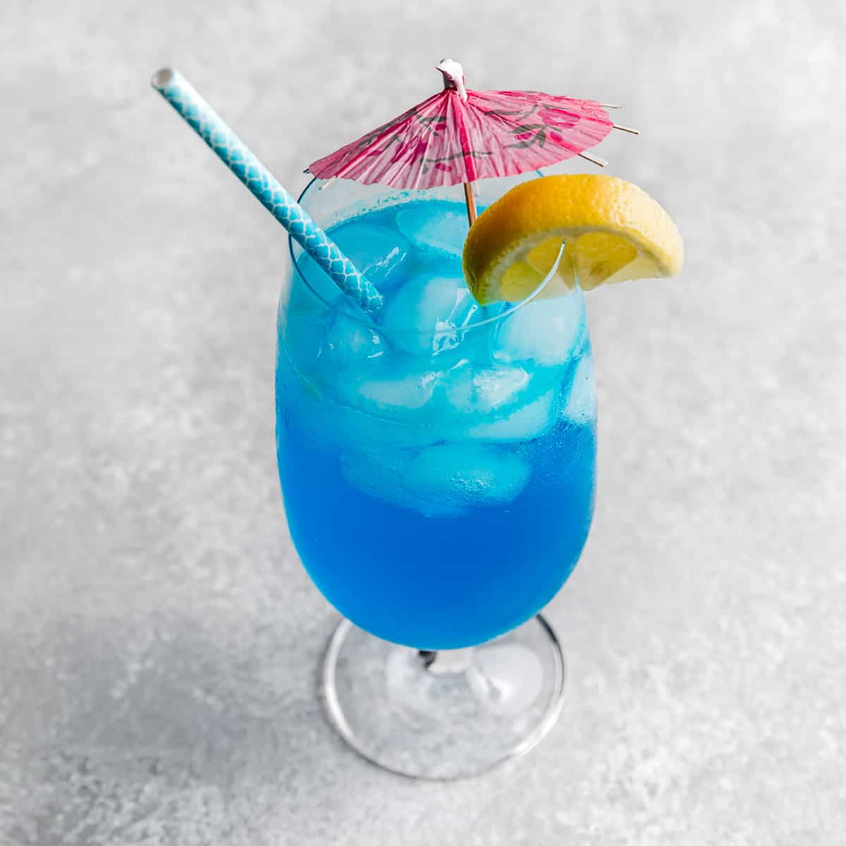A Blue Lagoon Mocktail in a large glass cup with a pink umbrella and lemon slice on the rim.