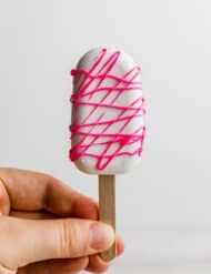 A hand holding a white chocolate covered Cakesicle that has been drizzled with hot pink melted chocolate.