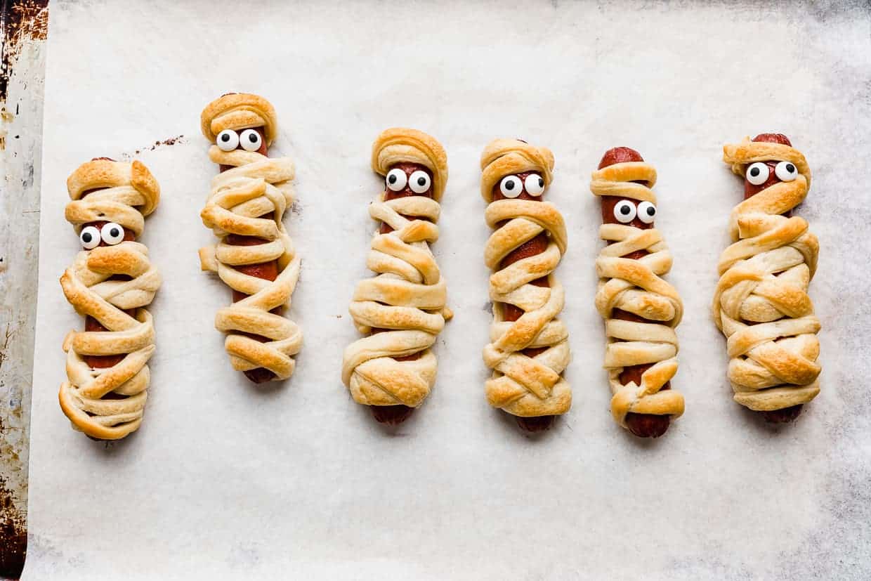 Mummy Hot Dogs on a white parchment lined baking sheet, with candy eyes on the mummy dogs.