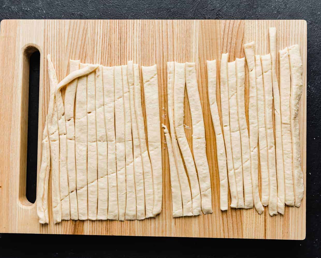 A wooden cutting board with a crescent roll sheet cut into strips on it.