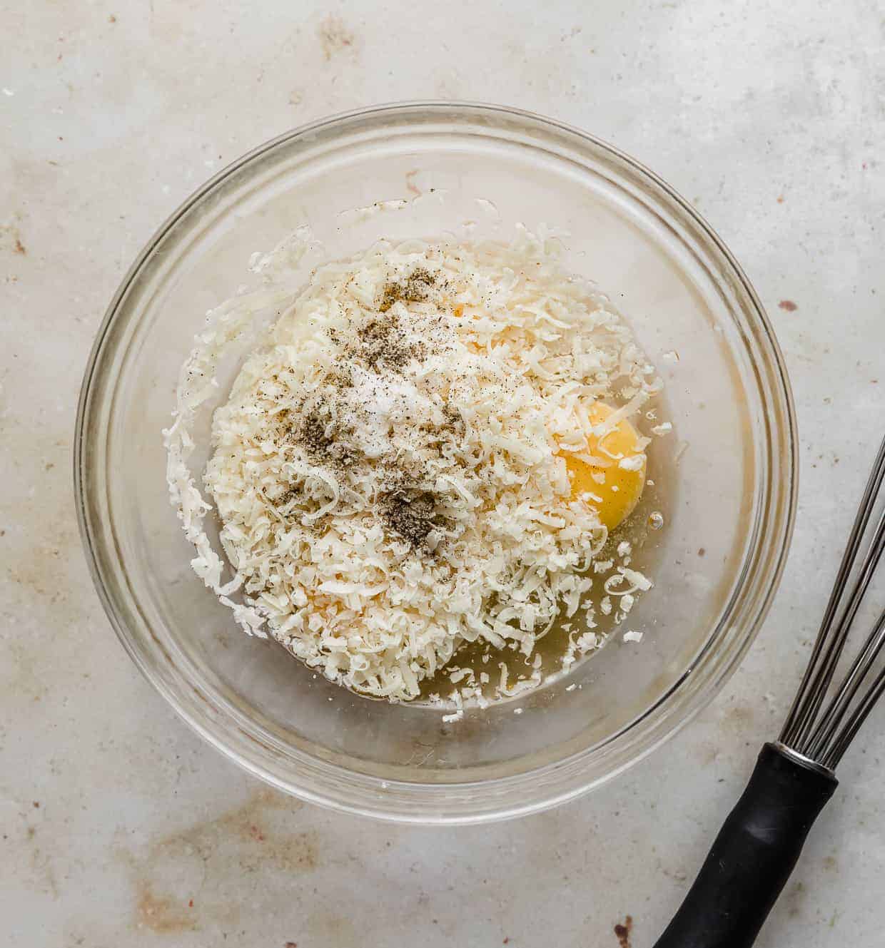 A glass bowl filled with grated parmesan, cracked eggs, and seasonings.