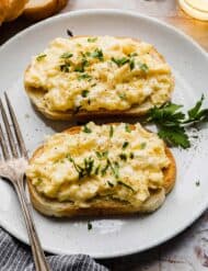 Two slices of bread topped with scrambled eggs and garnishes of chopped parsley.
