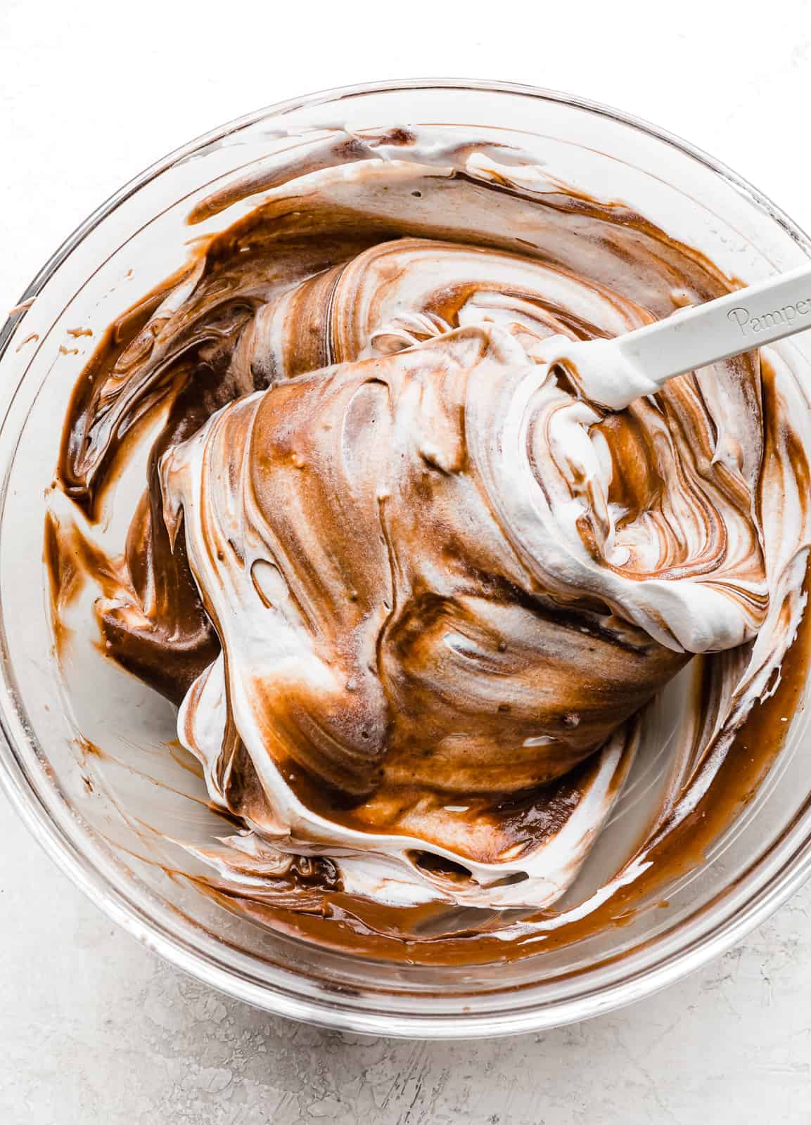 A spatula folding whipped cream into chocolate pudding in a large glass bowl.