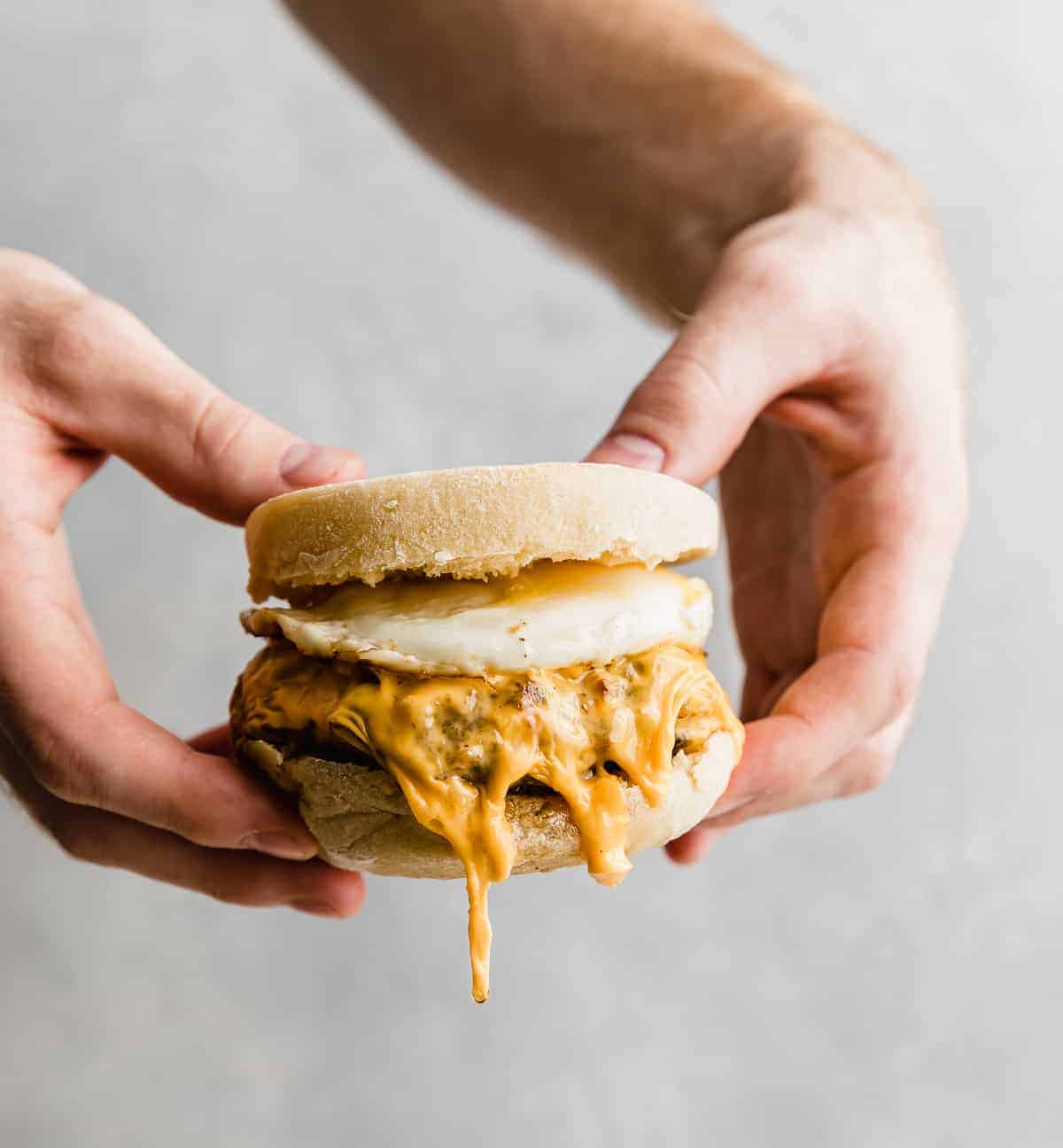 A set of hands holding a Homemade Sausage and Egg McMuffin against a gray background.