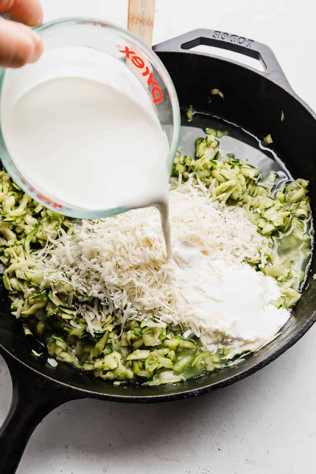 Heavy cream being poured overtop shredded zucchini that's in a black skillet.