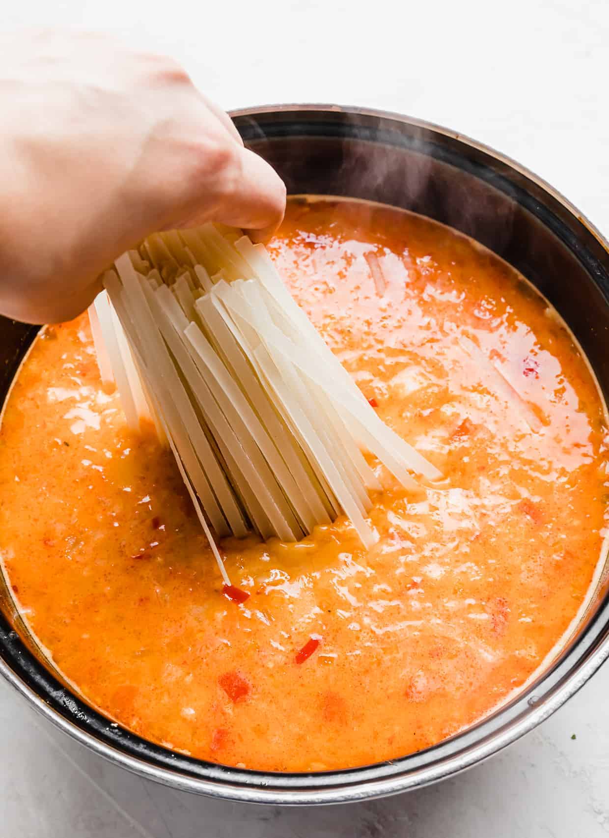 A hand setting linguini rice noodles into a pot of Thai Red Curry Noodle Soup.