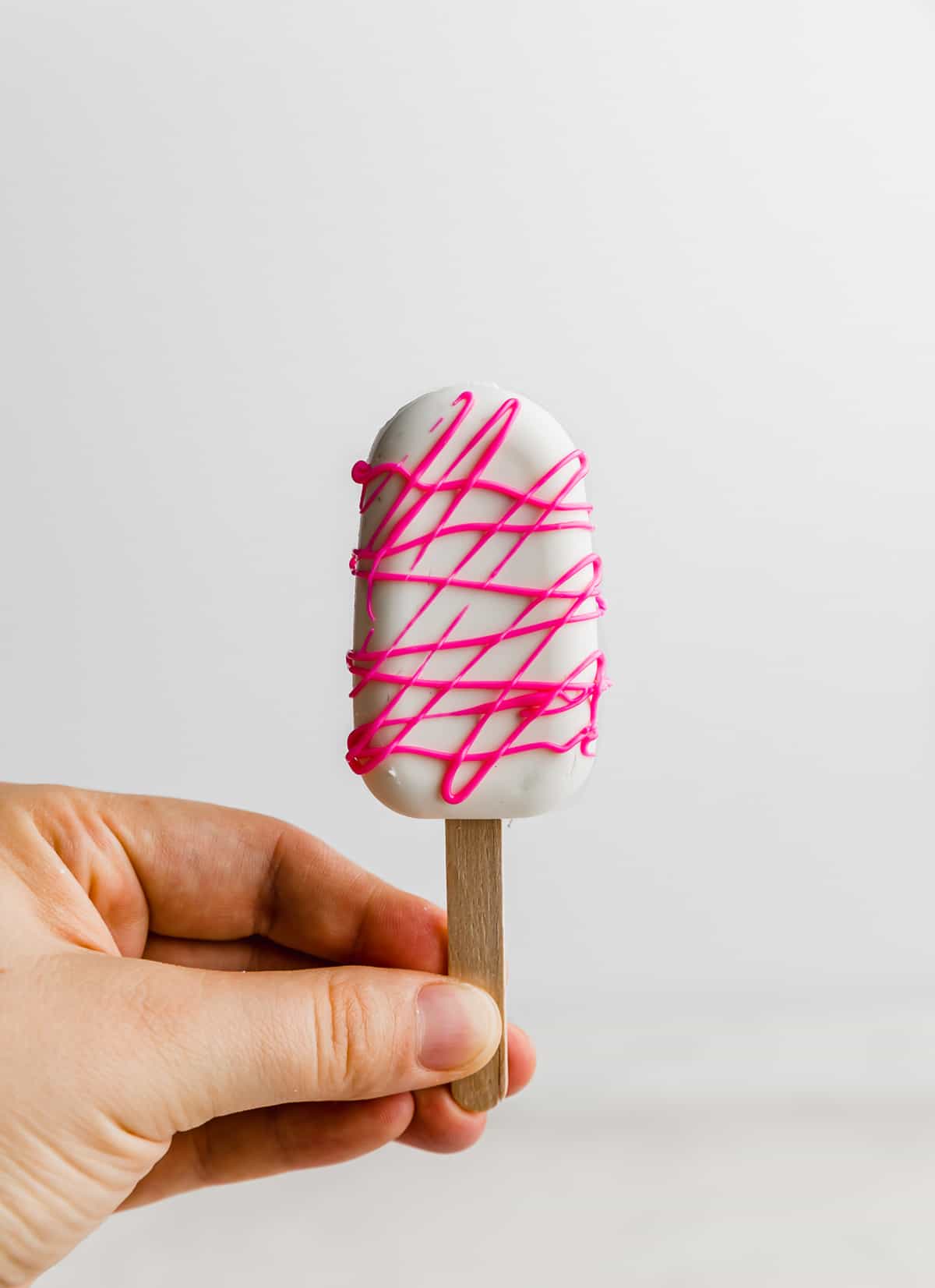 A hand holding up a white chocolate covered Cakesicle drizzled with hot pink chocolate.