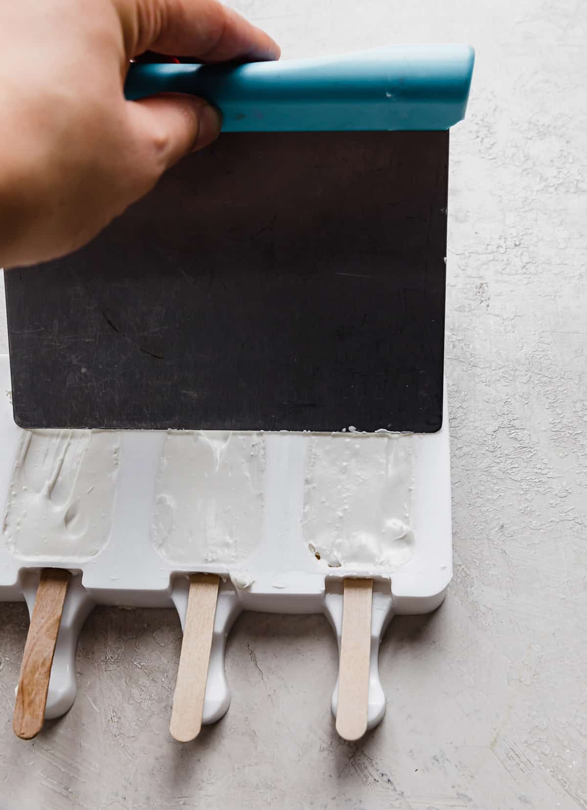 A metal scraper scraping off excess white chocolate from a white Cakesicle mold.