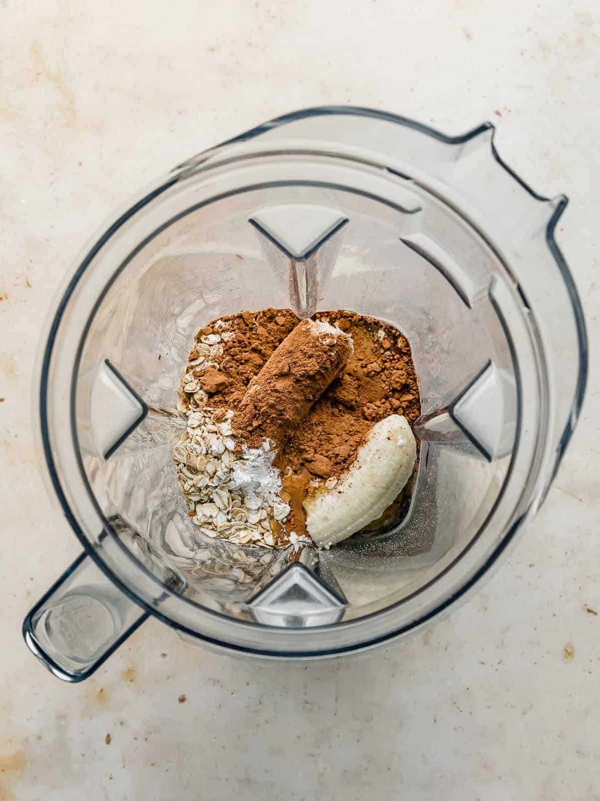 An overhead photo of a blender with a banana, oats, and cocoa unblended.