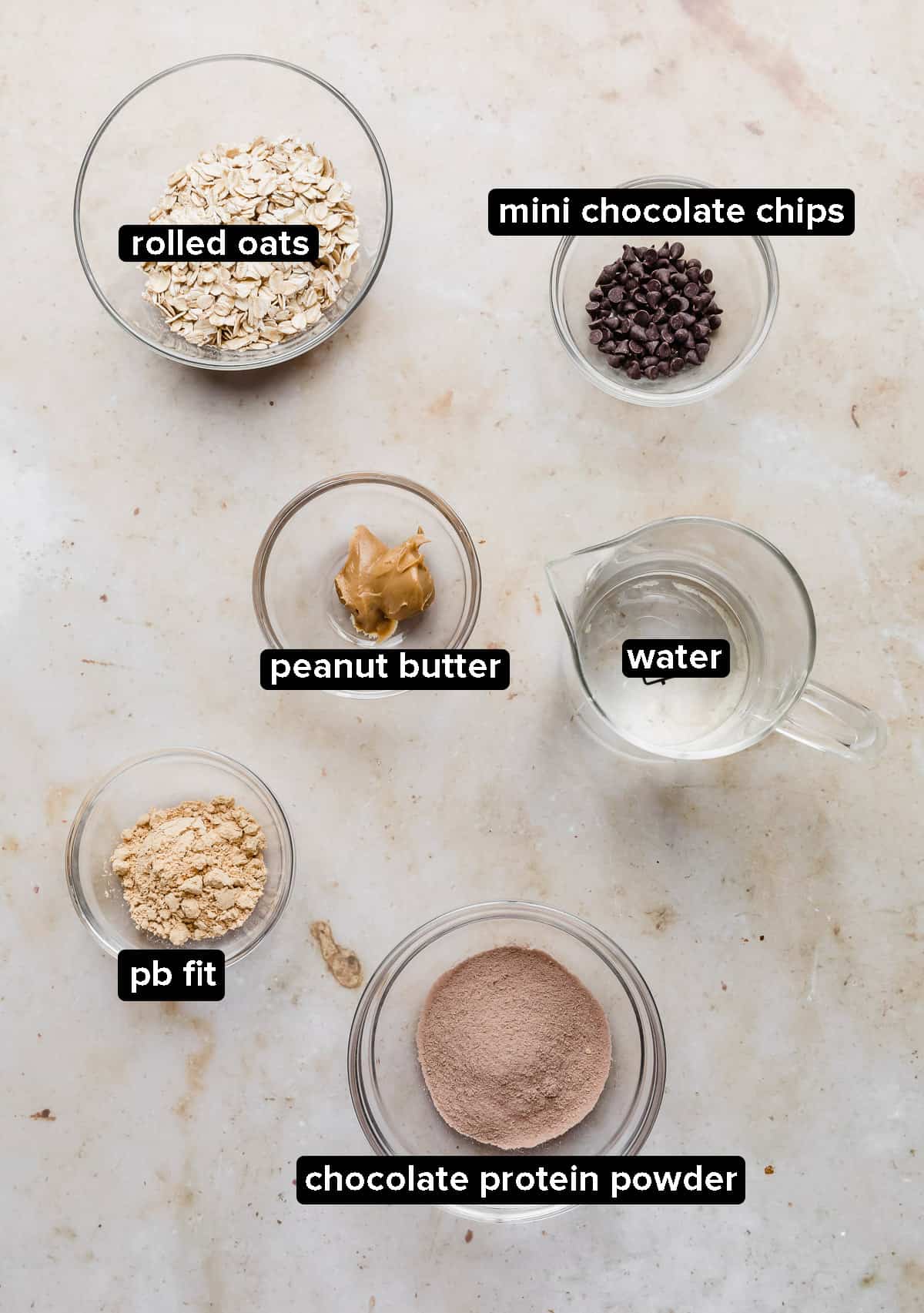 Chocolate Peanut Butter Protein Oatmeal ingredients in glass bowls on a tan marble background.