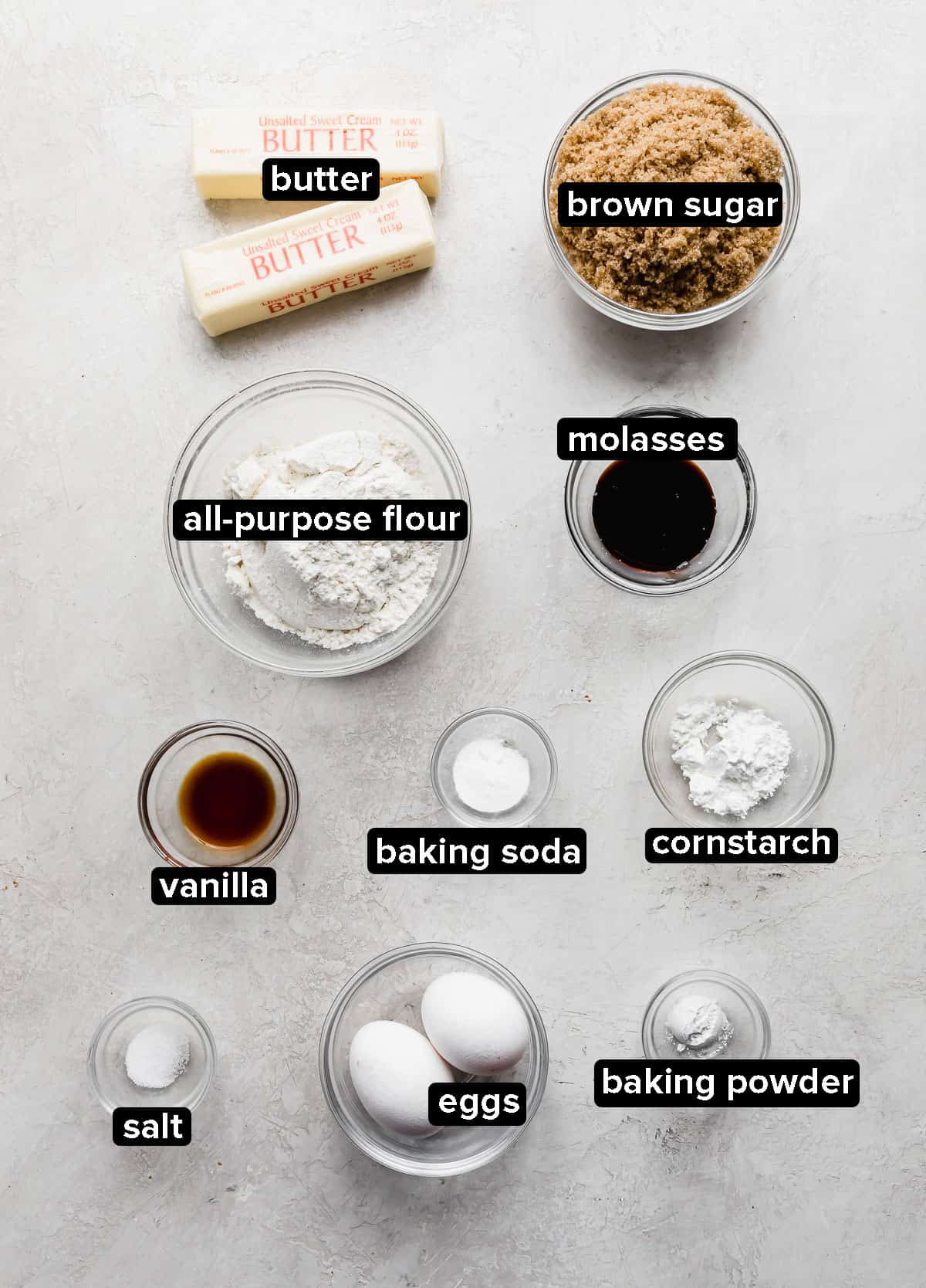 Ingredients in glass bowls such as: flour, butter, sugar, vanilla, salt, baking powder, baking soda, on a gray background.