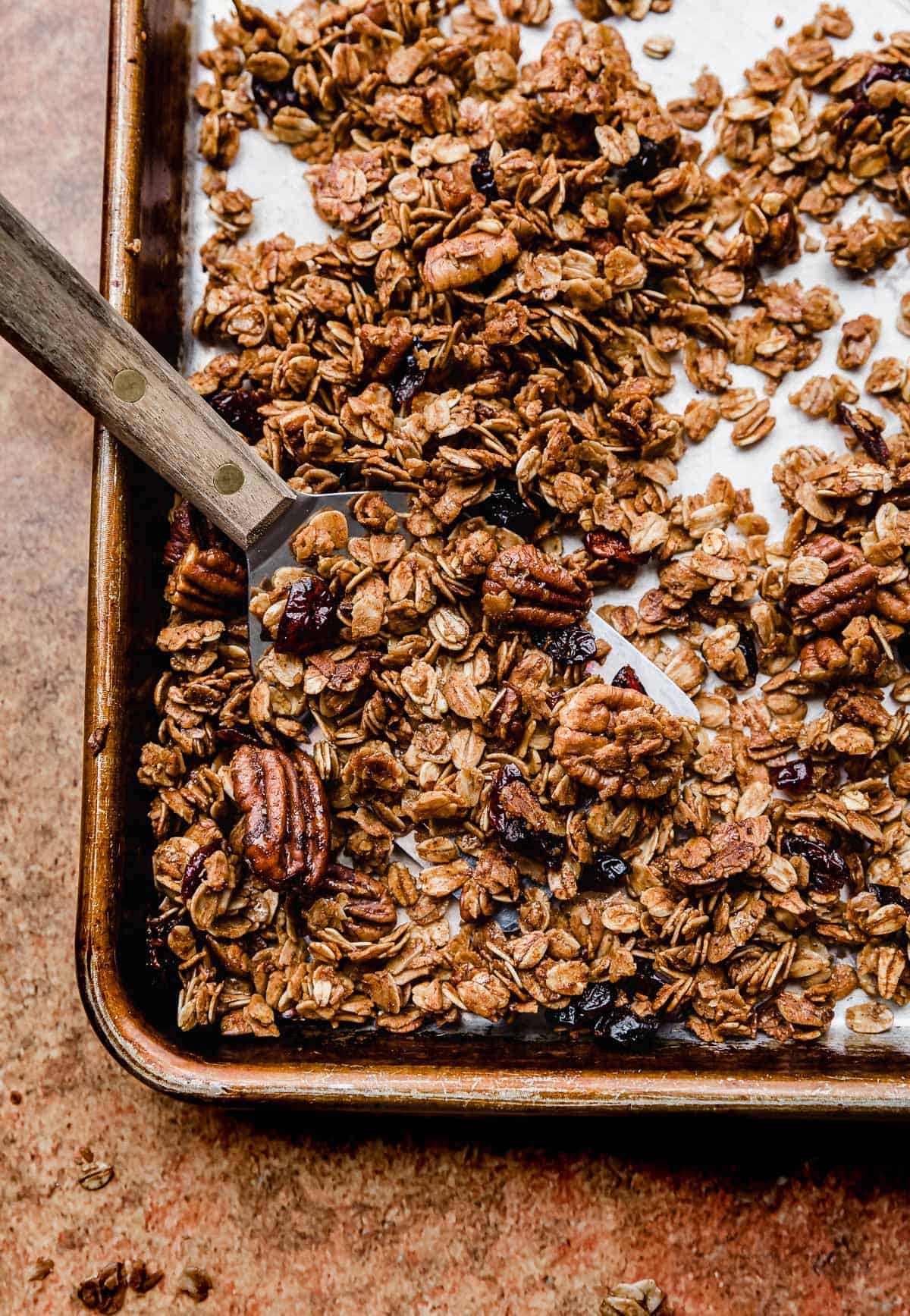 A metal spatula scooping up a deep amber colored gingerbread granola from a baking sheet.