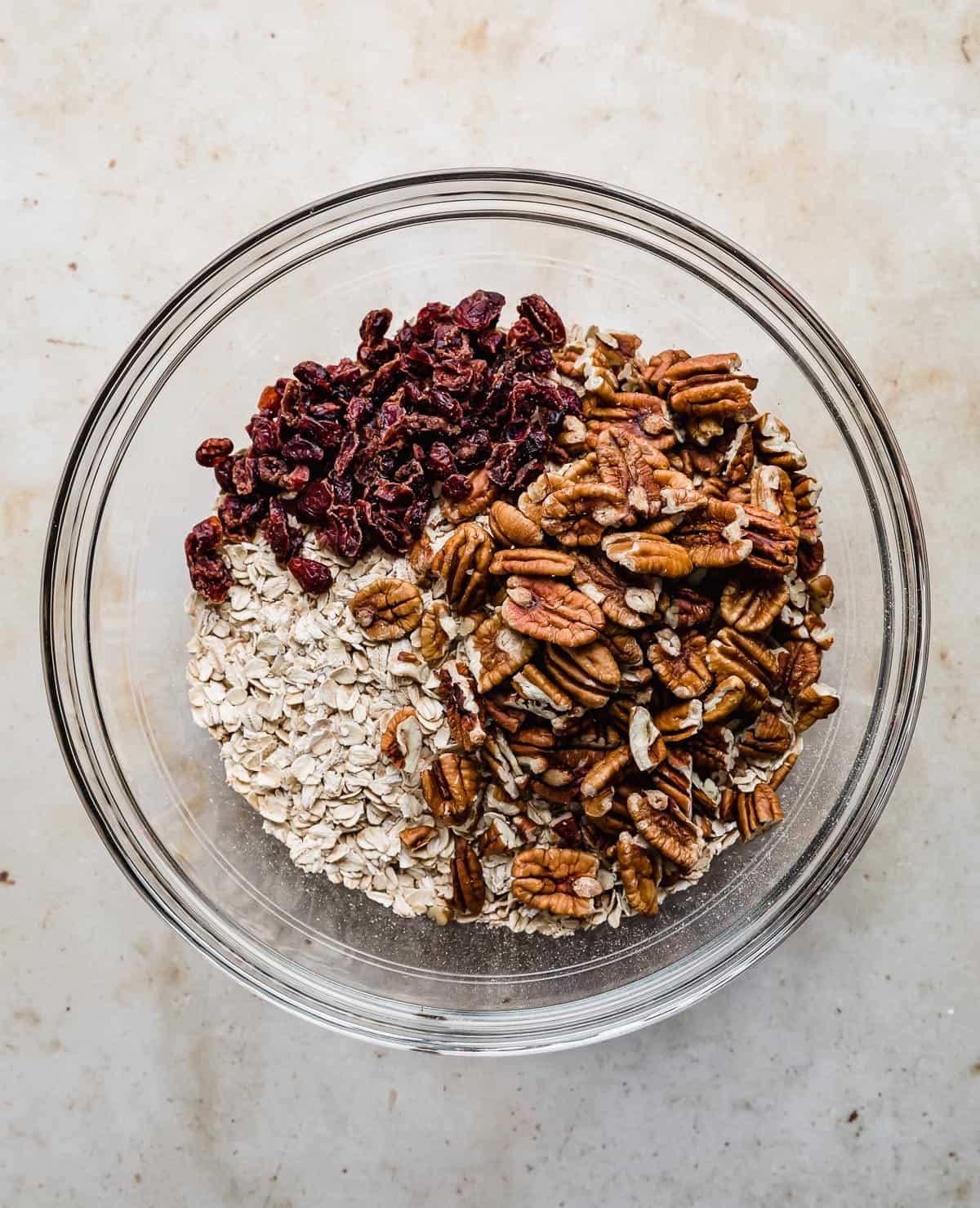 A glass bowl with Gingerbread Granola ingredients in it: pecans, dried cranberries, oats.