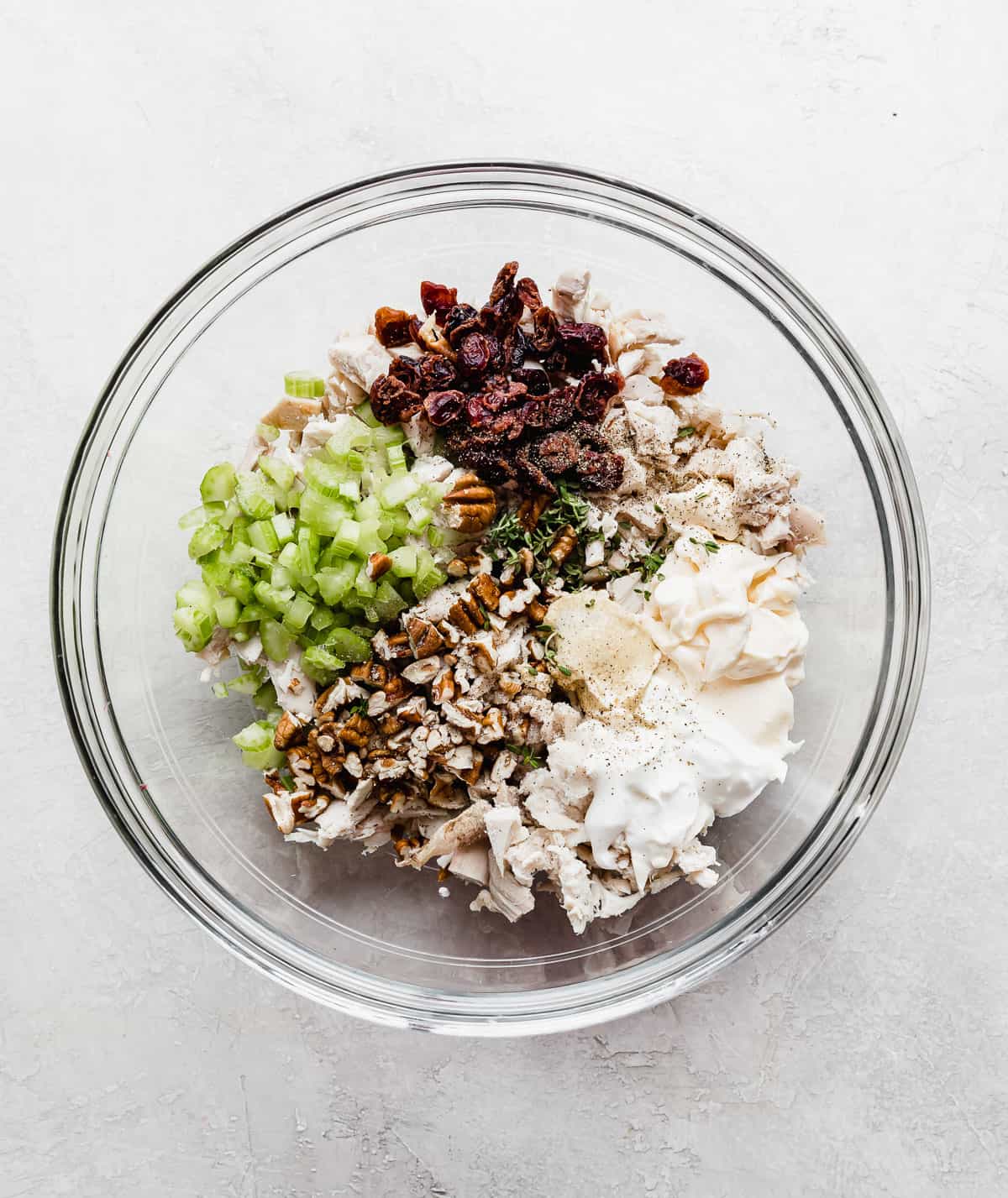 A glass bowl with turkey salad ingredients in it: celery, turkey, dried cranberries, pecans, mayo.