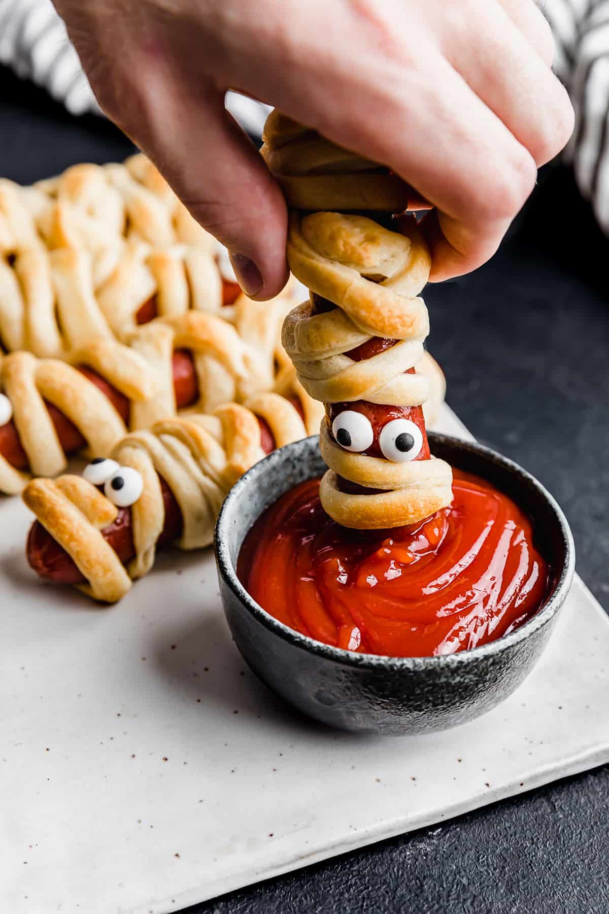A hand dipping a Mummy Hot Dog into a bowl of ketchup.