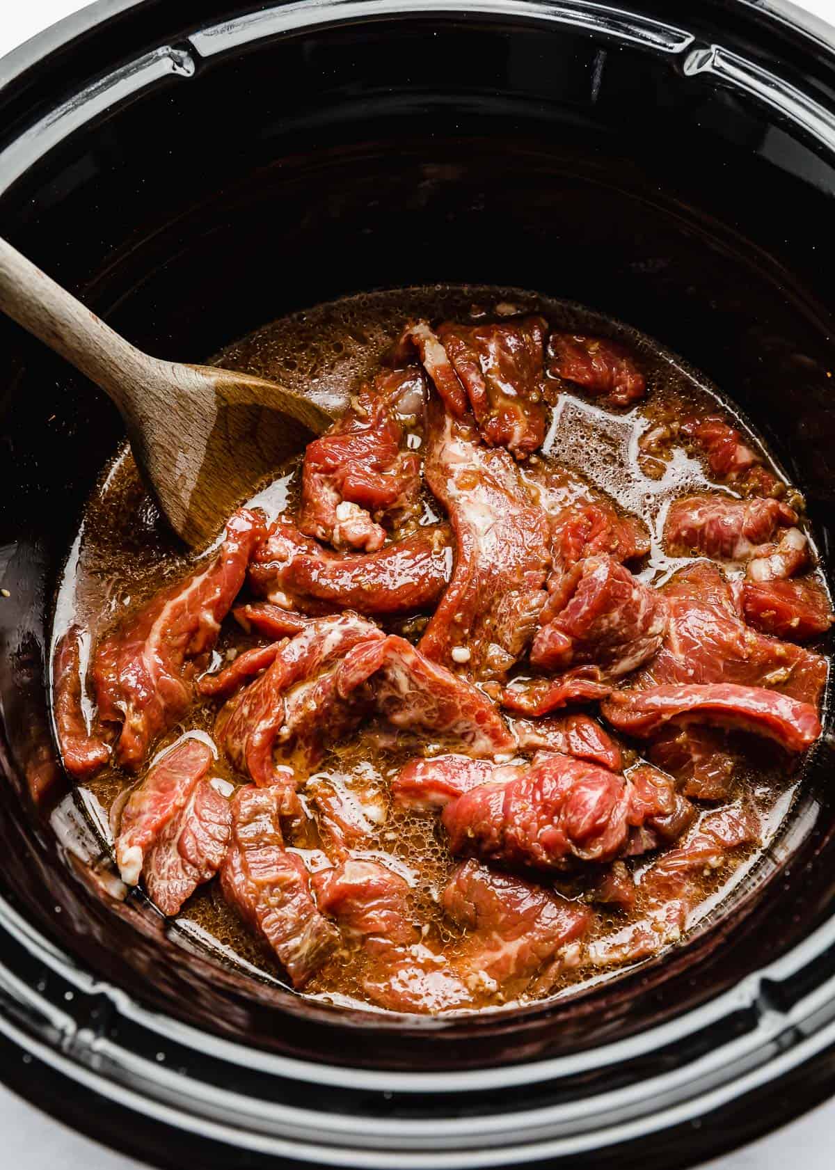 Raw flank steak strips in a slow cooker covered in a brown juice.