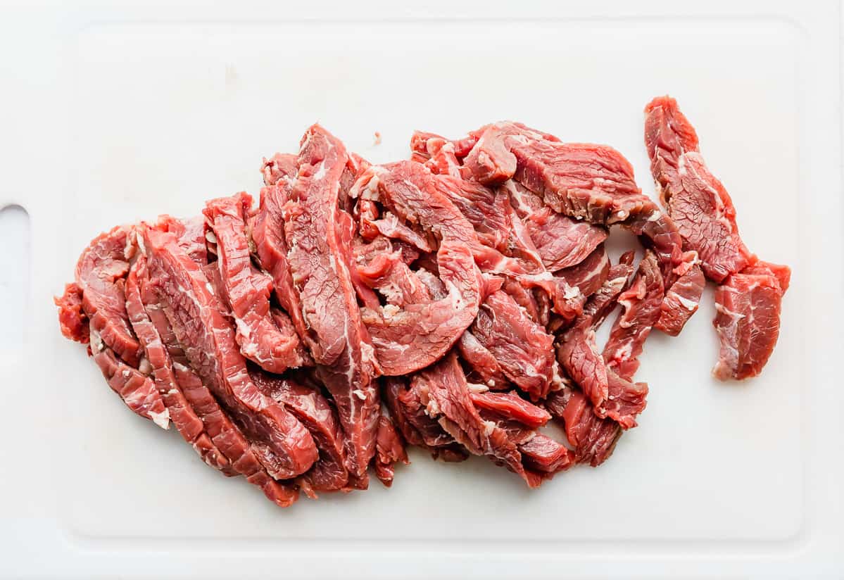 Flank steaks cut into thin strips on a white cutting board.