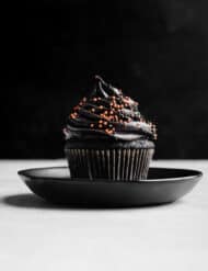 A single Black Velvet Cupcake topped with a black frosting and orange and black sprinkles against a black background.