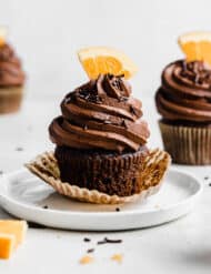 A chocolate frosting topped chocolate orange cupcake on a white plate, with a fresh slice of an orange as a topper.