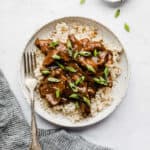 A white plate with Slow Cooker Mongolian Beef topped with sliced green onions on brown rice.