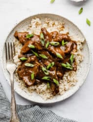 A white plate with Slow Cooker Mongolian Beef topped with sliced green onions on brown rice.