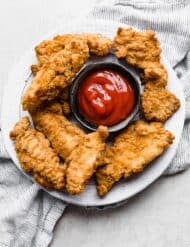 Air Fryer Frozen Chicken Tenders on a white plate with a black bowl in the center full of red ketchup.