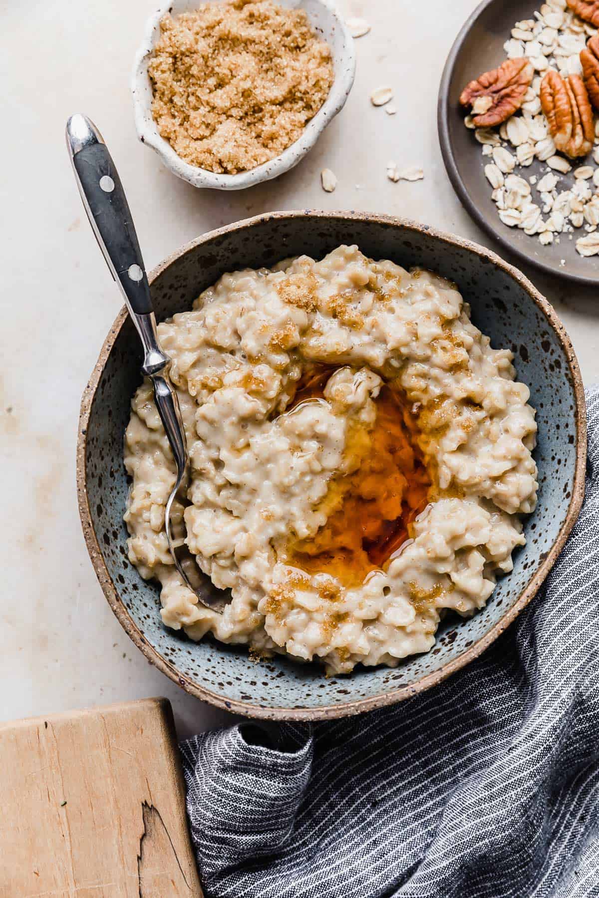 A bowl of Maple and Brown Sugar Oatmeal on a cream background with a bowl of pecans and brown sugar to the side.