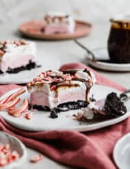 A square slice of Christmas Ice Cream Cake (Oreo crust, pink peppermint ice cream, whipped topping, hot fudge, and crushed candy canes).