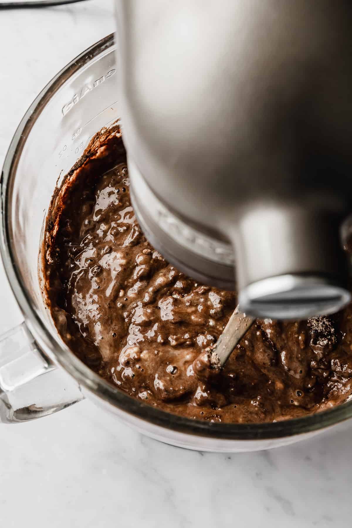 A dark chocolate Bundt Cake batter being mixed in a glass stand mixer bowl attached to a silver kitchen aid.