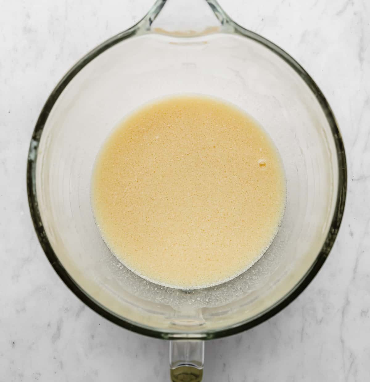 A light yellow colored liquid mixture in a glass mixing bowl on a white marble background.