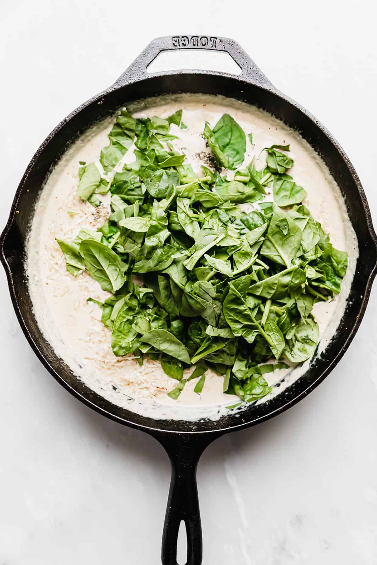 Spinach overtop a tan tuscan chicken gravy mixture in a skillet on a white background.