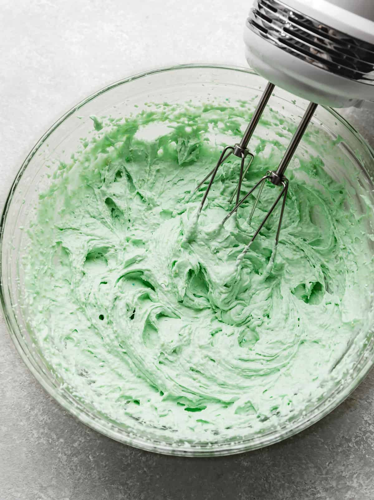 A hand mixer beating together a mint colored mousse to frost Crumbl Mint Chip Ice Cream Cookies.