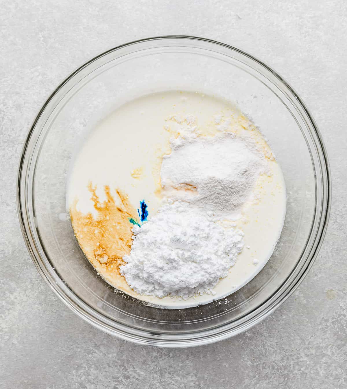 A glass bowl with heavy cream, powdered sugar, vanilla pudding powder, and blue food color drops in it.