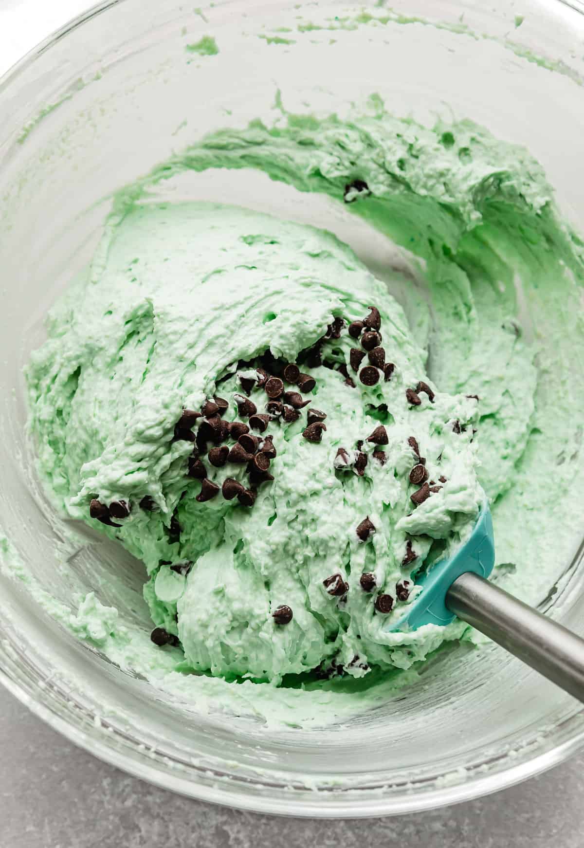 Close up photo of a spatula folding mini chocolate chips into a mint green colored mint mousse.