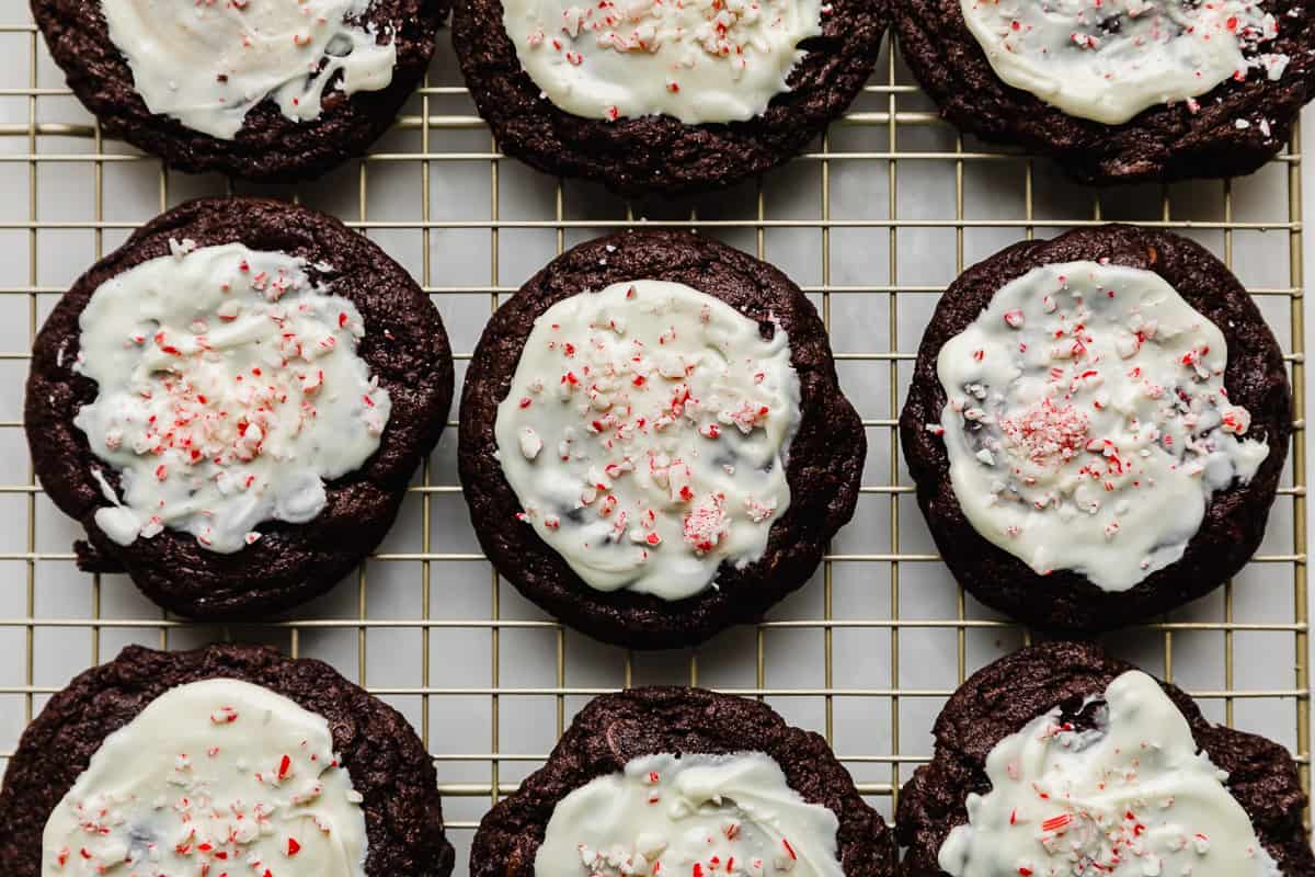 Chocolate peppermint cookies topped with white chocolate and candy canes on a golden wire cooling rack.