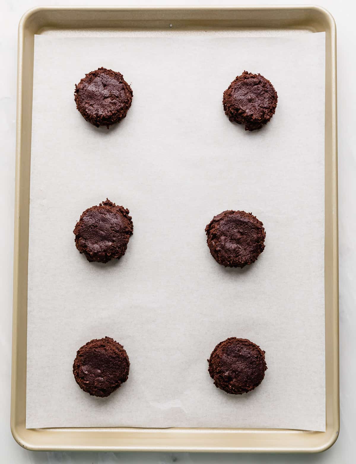 Six dark chocolate cookie dough balls on a white parchment lined baking sheet.