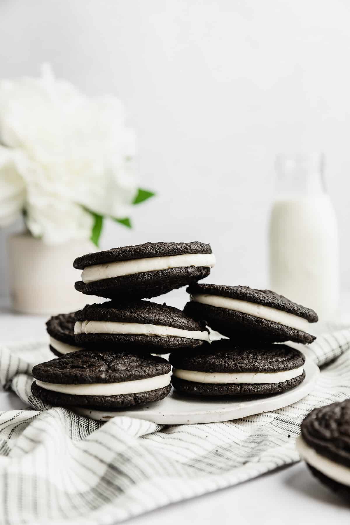 Homemade Oreo Cookies stacked on a white plate with a glass of milk and a white flower in the background.