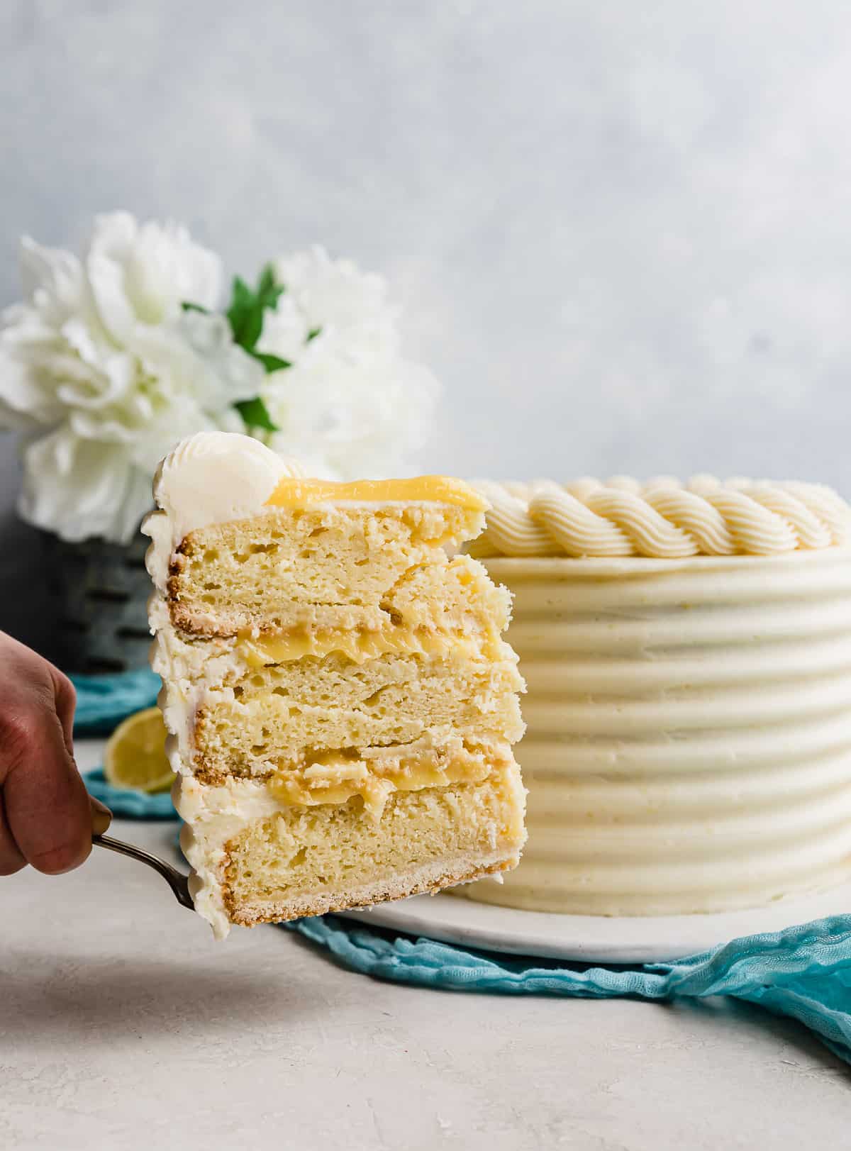 A slice of lemon bar layer cake being held up in front of the full cake.