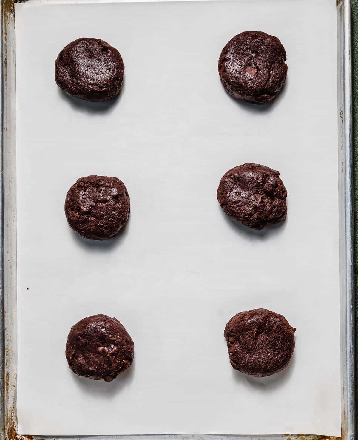 Six chocolate cookie dough balls on a white parchment lined baking sheet.
