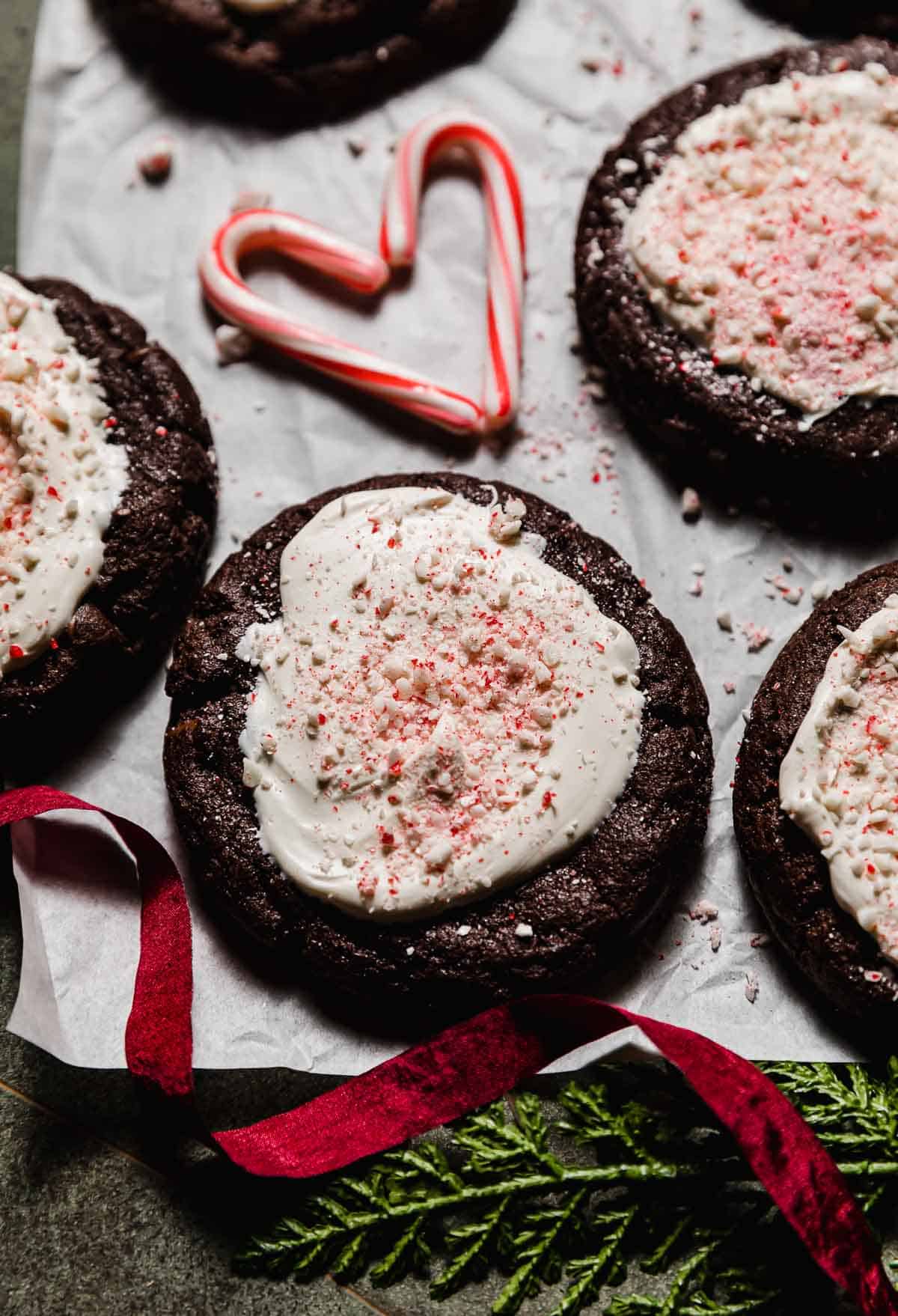 Chocolate cookie topped with white chocolate and crushed candy canes (Peppermint Bark Cookie) on a white parchment paper with a small candy cane heart in the background.